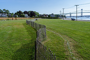 Wooden beach fencing runs along a grassy roadside lawn, with houses, bridge, and calm bay water in background.